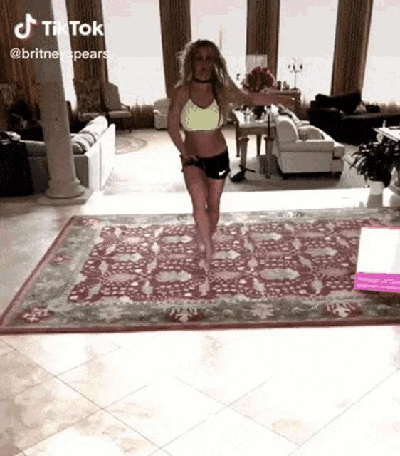 a woman in a yellow top and black shorts is dancing on a rug in a living room .