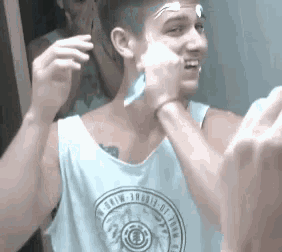 a man in a tank top is shaving his face in front of a mirror while a woman looks on .