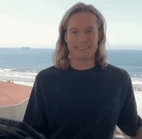 a man with long blonde hair wearing a black shirt is smiling in front of the ocean