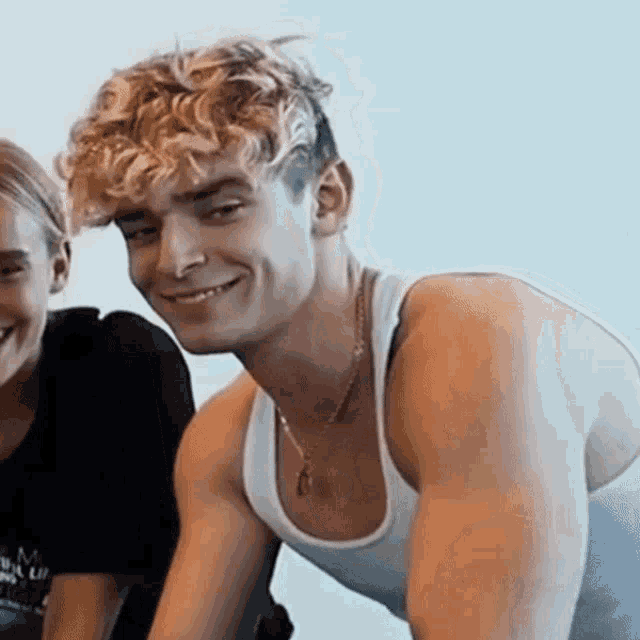a man with curly hair wearing a white tank top and a necklace smiles