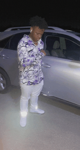 a man in a purple shirt and white pants is standing next to a silver car