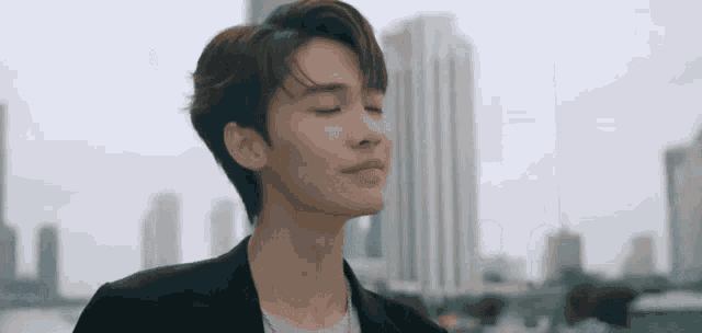 a young man in a black suit is standing in front of a city skyline with his eyes closed .