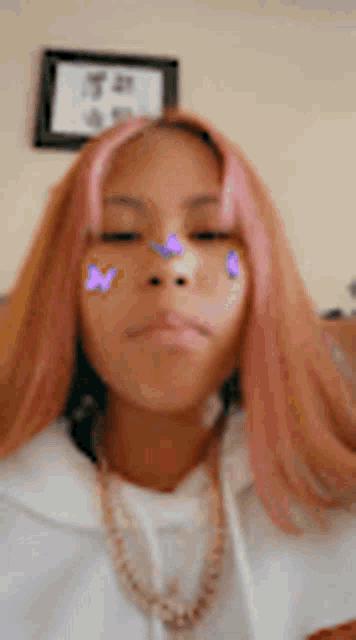 a woman with pink hair and purple butterflies on her face is wearing a white shirt and a gold chain .