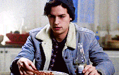 a young man is sitting at a table eating a bowl of food while holding a bottle of beer .
