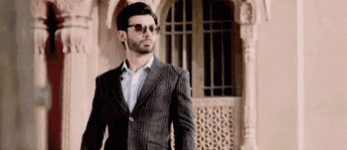 a man wearing sunglasses and a suit is walking in front of a building .