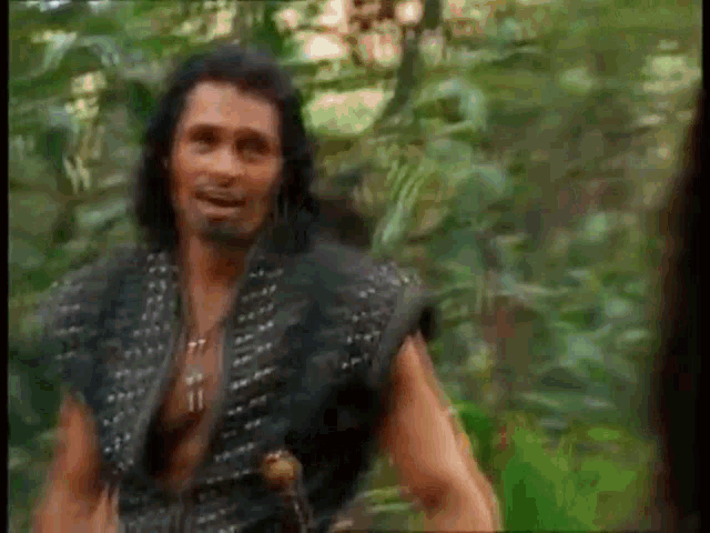 a man with long black hair and a beard is standing in the woods .