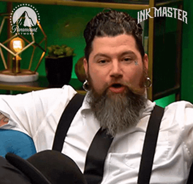 a man with a beard and suspenders is sitting in a chair with his mouth open .