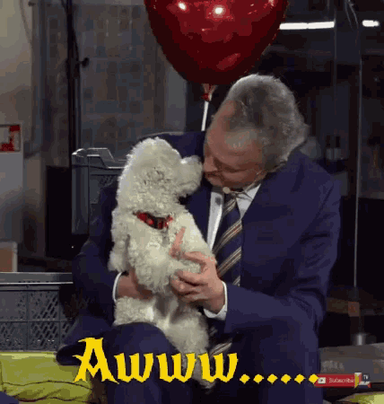 a man in a suit and tie is holding a small white dog with a heart shaped balloon behind him that says awww