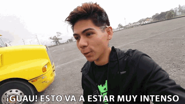a man in a black jacket stands in front of a yellow truck with the words guau esto va a estar muy intenso on the bottom