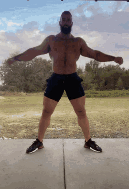 a shirtless man with a beard stands on a sidewalk with his arms outstretched