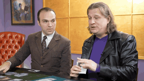 two men are sitting at a table with cards on it and one of them is wearing a purple shirt