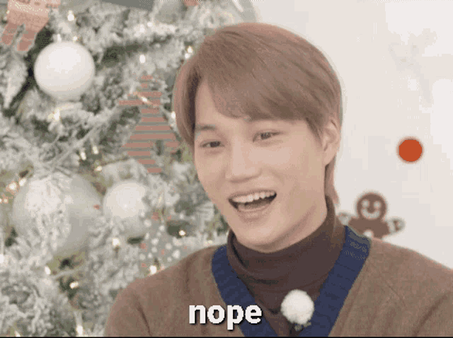 a young man is smiling in front of a christmas tree and the word nope is on the bottom of his face