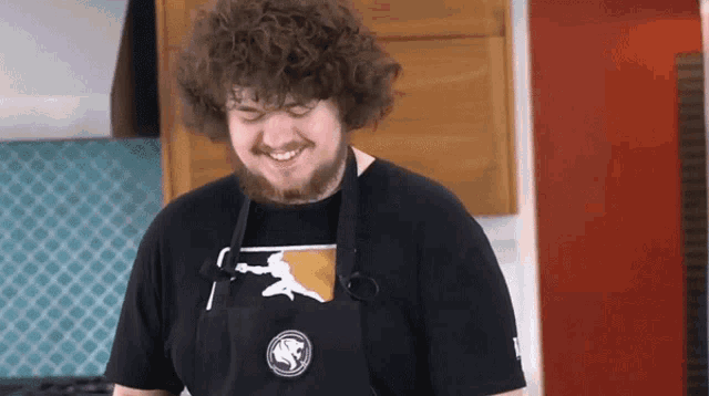 a man with curly hair wearing an apron and a t-shirt with a lion on it smiles