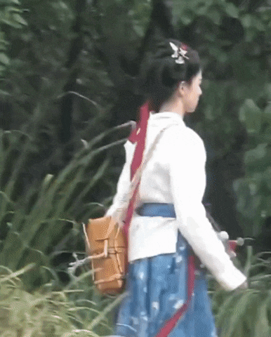 a woman in a blue skirt and white jacket is walking through the woods carrying a yellow bag .