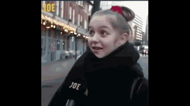 a young girl wearing a black scarf and a red bow in her hair is standing on a street .
