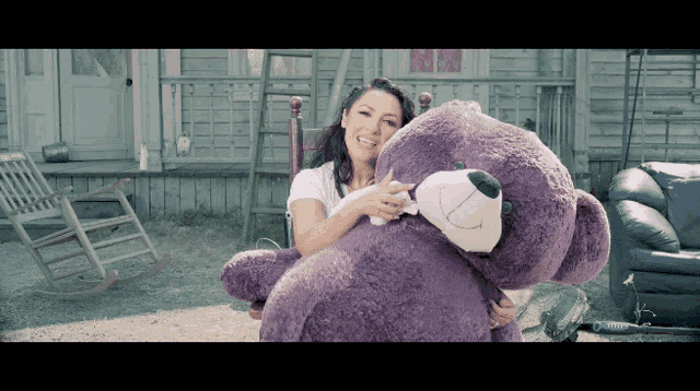 a woman is holding a purple teddy bear in front of a house