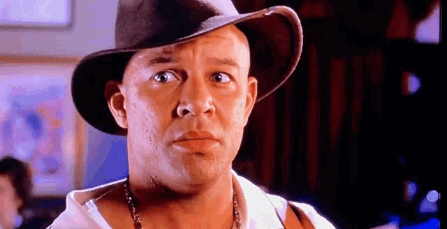a bald man wearing a cowboy hat and a necklace looks at the camera