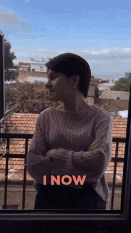 a woman in a pink sweater is standing in front of a window with the words " i now " written in red