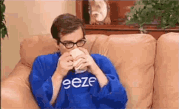 a man is sitting on a couch drinking from a cup while wearing a blue shirt that says eeze .