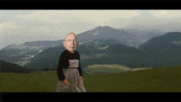 a man in a black shirt and apron is standing on a hill with mountains in the background