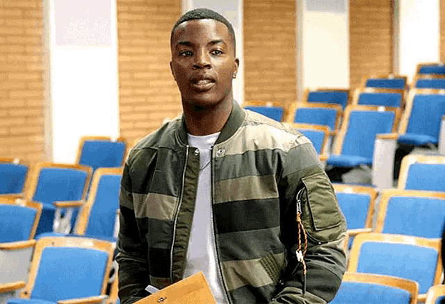 a young man in a bomber jacket is standing in a lecture hall holding a briefcase .