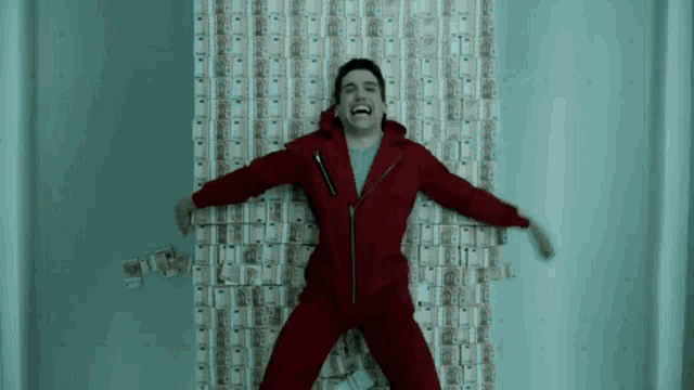 a man in a red suit is laying on a wall covered in money and laughing .