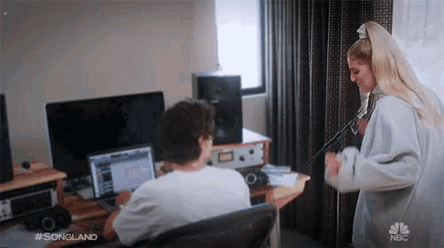 a woman is singing into a microphone while a man sits at a desk with a laptop on it