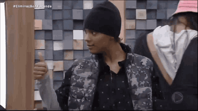 a man wearing a black beanie and a camouflage vest is giving a thumbs up
