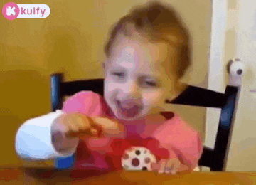 a little girl is sitting at a table eating a cookie with her mouth open .