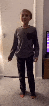 a young boy in a gray shirt and black pants is standing in front of a television .