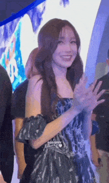 a woman in a black dress is smiling and waving at the camera while standing in a crowd .