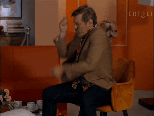 a man in a suit and tie is sitting in a chair in front of an orange wall that says ert