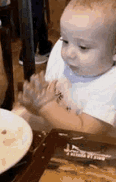 a baby in a bib is sitting at a table with a plate of food ..