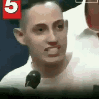 a man in a white shirt is talking into a microphone with the number 5 in the background .