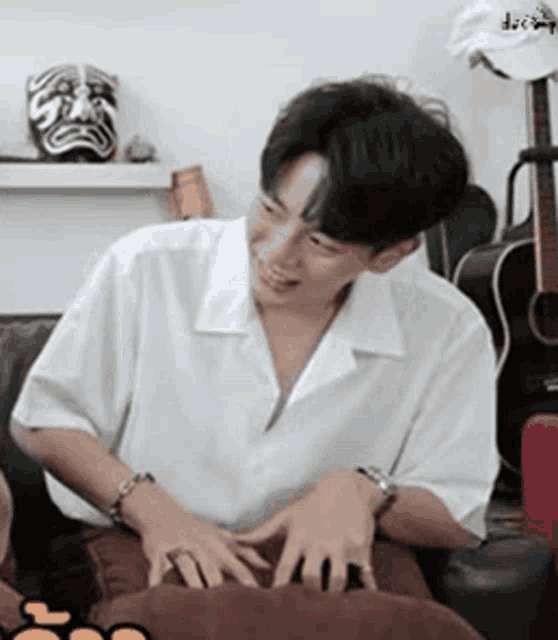a young man in a white shirt is sitting on a couch with his hands on a pillow .