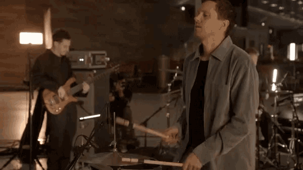 a man playing drums in a band with a man playing guitar in the background