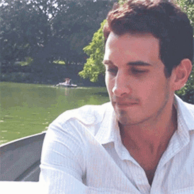 a man in a white shirt sits in front of a lake with a boat in the background