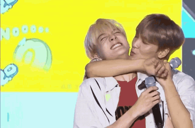 two young men are hugging each other while holding microphones in front of a yellow background .
