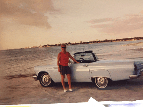 a woman stands in front of a white thunderbird on a beach