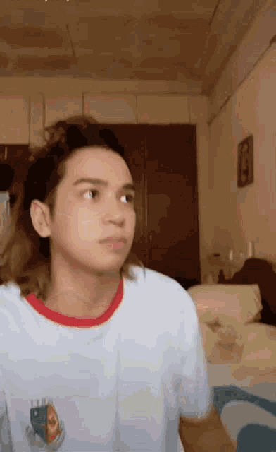 a young man with long hair is wearing a white t-shirt with a red collar and is standing in a bedroom .