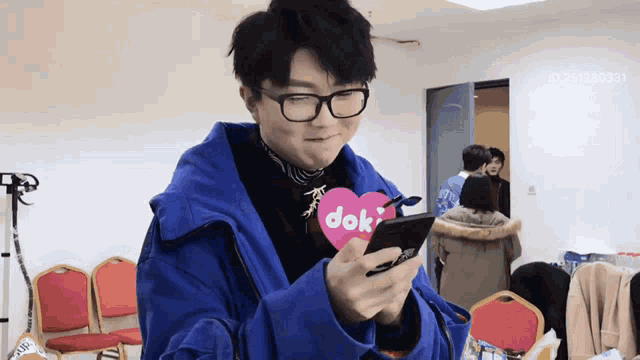 a man wearing glasses and a blue jacket is holding a cell phone and a heart that says dok on it