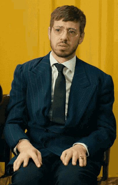 a man in a suit and tie is sitting with his hands folded in front of a yellow curtain