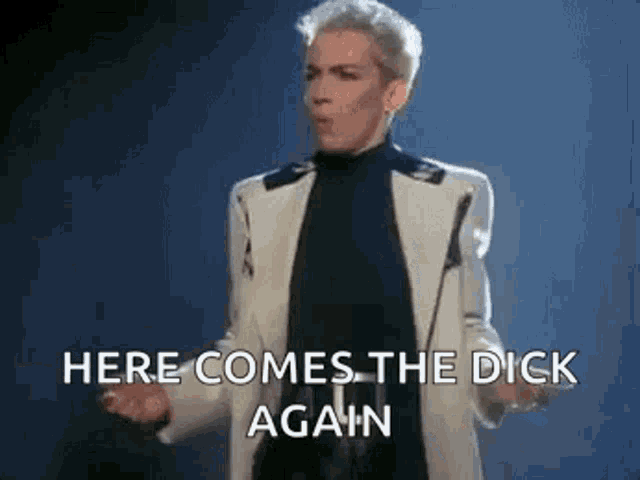 a man in a white suit and black turtleneck is standing in front of a microphone and saying `` here comes the dick again ''