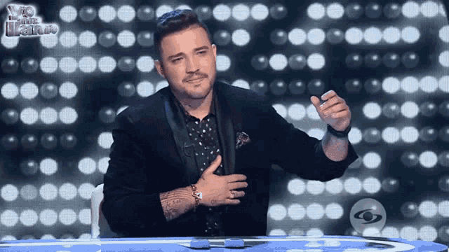 a man in a tuxedo is sitting at a table with his hand on his chest