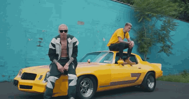 two men are sitting on the hood of a yellow car