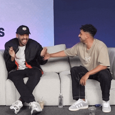 two men are sitting on a couch and one of them is wearing a bomber jacket