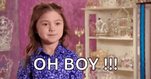a little girl in a purple shirt is standing in front of a shelf and saying `` oh boy !!! '' .