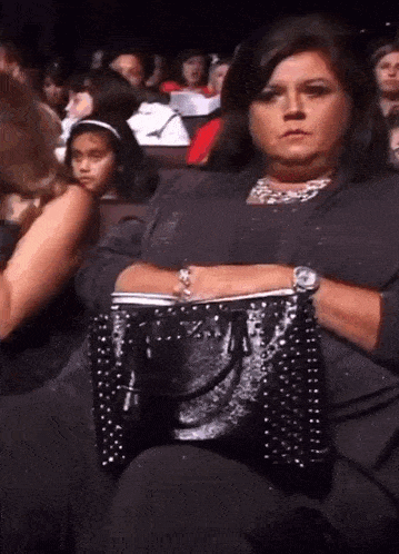 a woman is sitting in a theatre holding a black purse and wearing a watch .