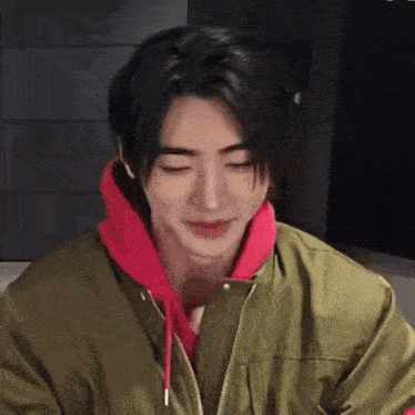 a young man wearing a green jacket and a red hoodie is smiling with his eyes closed .