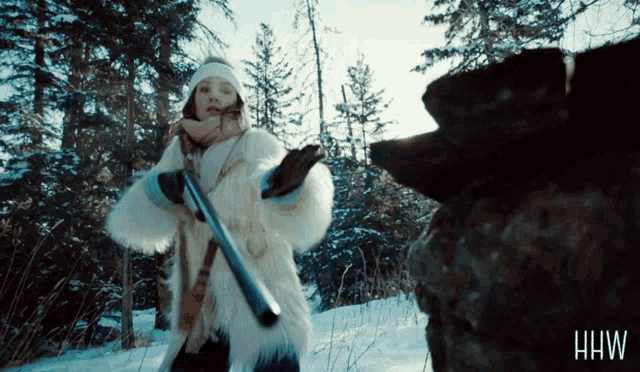 a woman in a fur coat holds a shotgun in front of a bear with the letters hw on the bottom right
